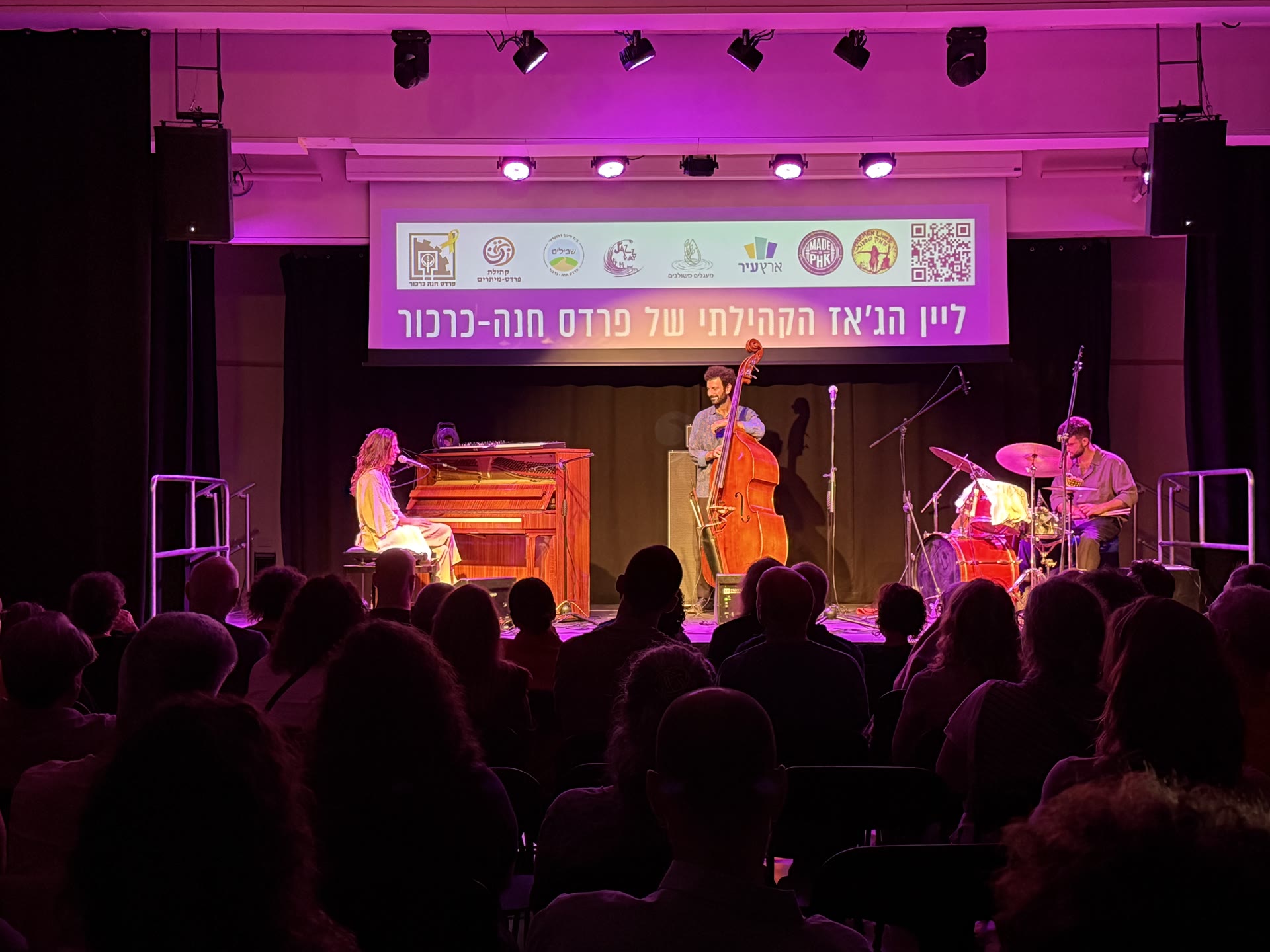 מופע ג׳אז חי בליין הקהילתי — פסנתר, קונטרבס ותופים על הבמה מול קהל, תחת שלט ליין הג׳אז הקהילתי